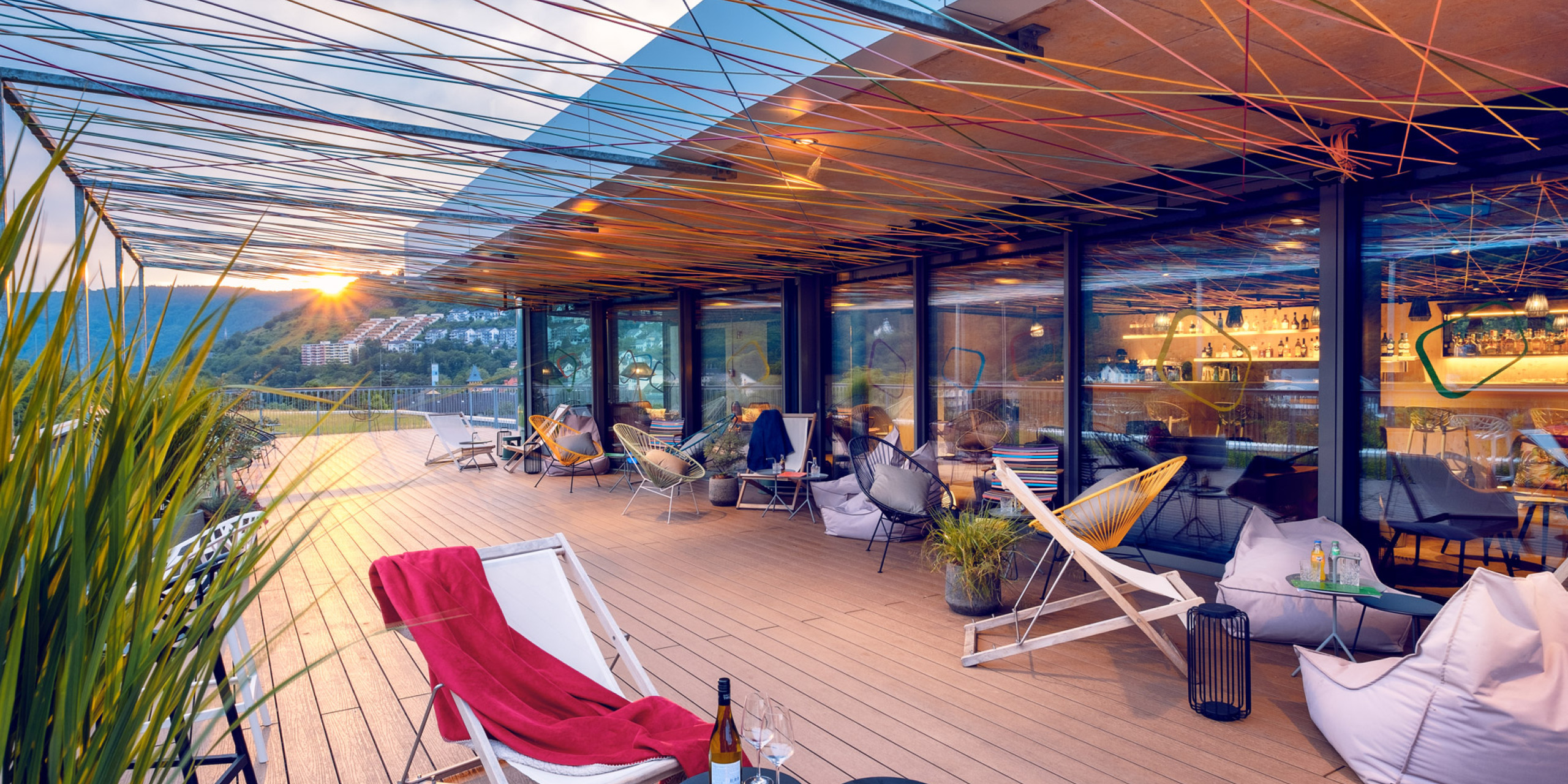 Gemütliche Terrasse mit modernem Design, geschmückt mit bunten Fäden. Sonnenuntergang im Hintergrund, Loungemöbel, Pflanzen und Getränke auf kleinen Tischen laden zum Entspannen ein.