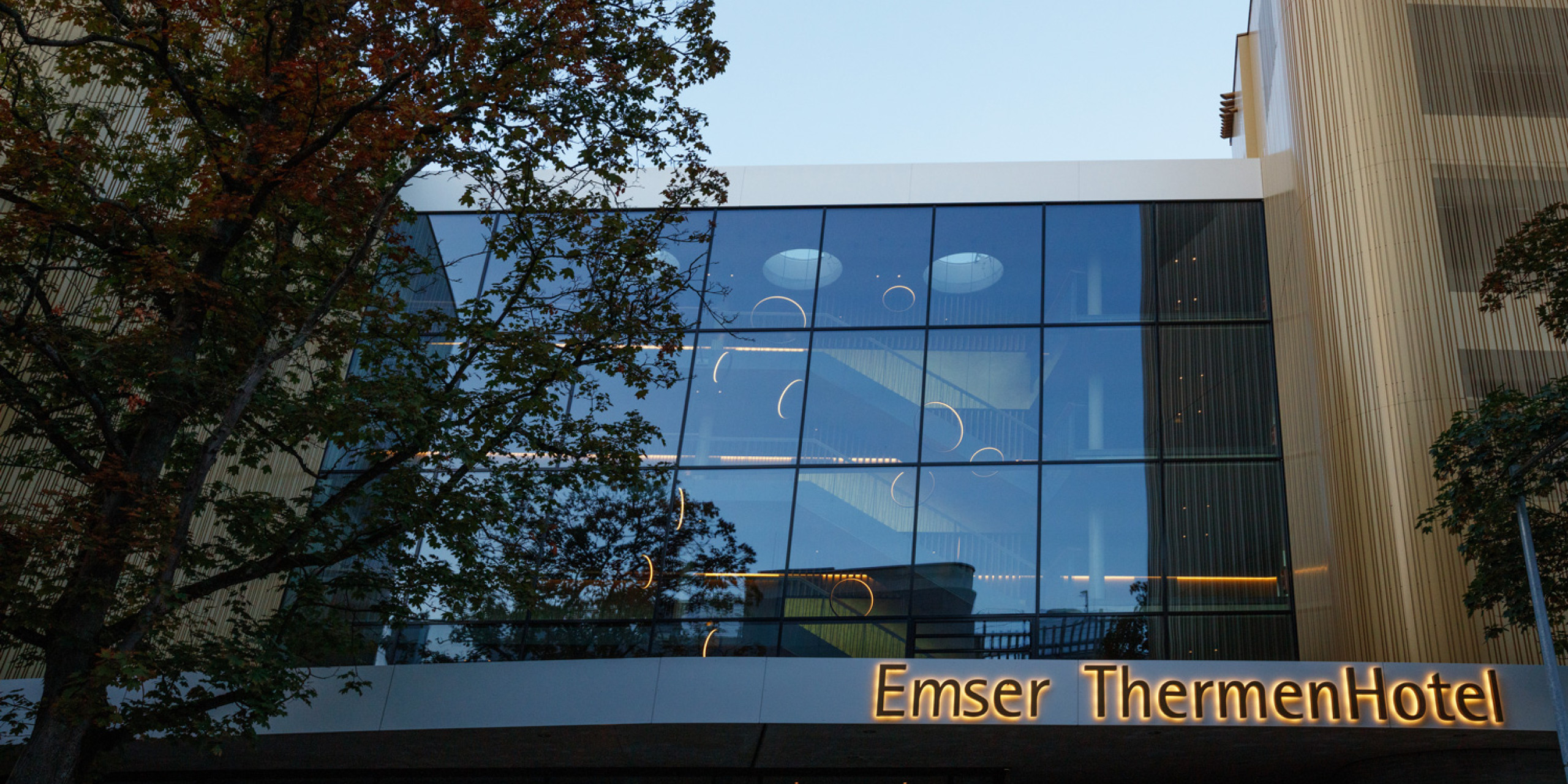 Moderne Architektur des Emser ThermenHotels mit großen Glasscheiben und stilvollen Lichtringen. Ein Herbstbaum im Vordergrund ergänzt die harmonische Szenerie unter blauem Himmel.