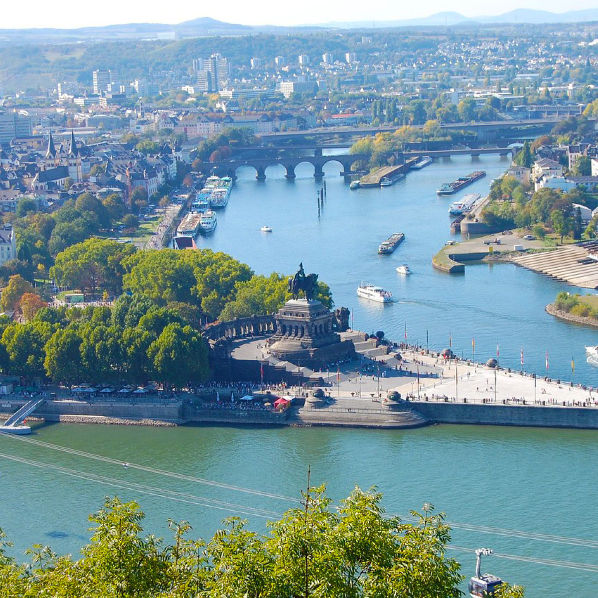 emser thermenhotel ausflugsziele koblenz Panoramablick über das Rheintal mit dem Deutschen Eck, wo Mosel und Rhein zusammenfließen. Grüne Bäume umrahmen die Ufer, zahlreiche Boote sind auf dem Wasser unterwegs, und die Stadt ist sichtbar.